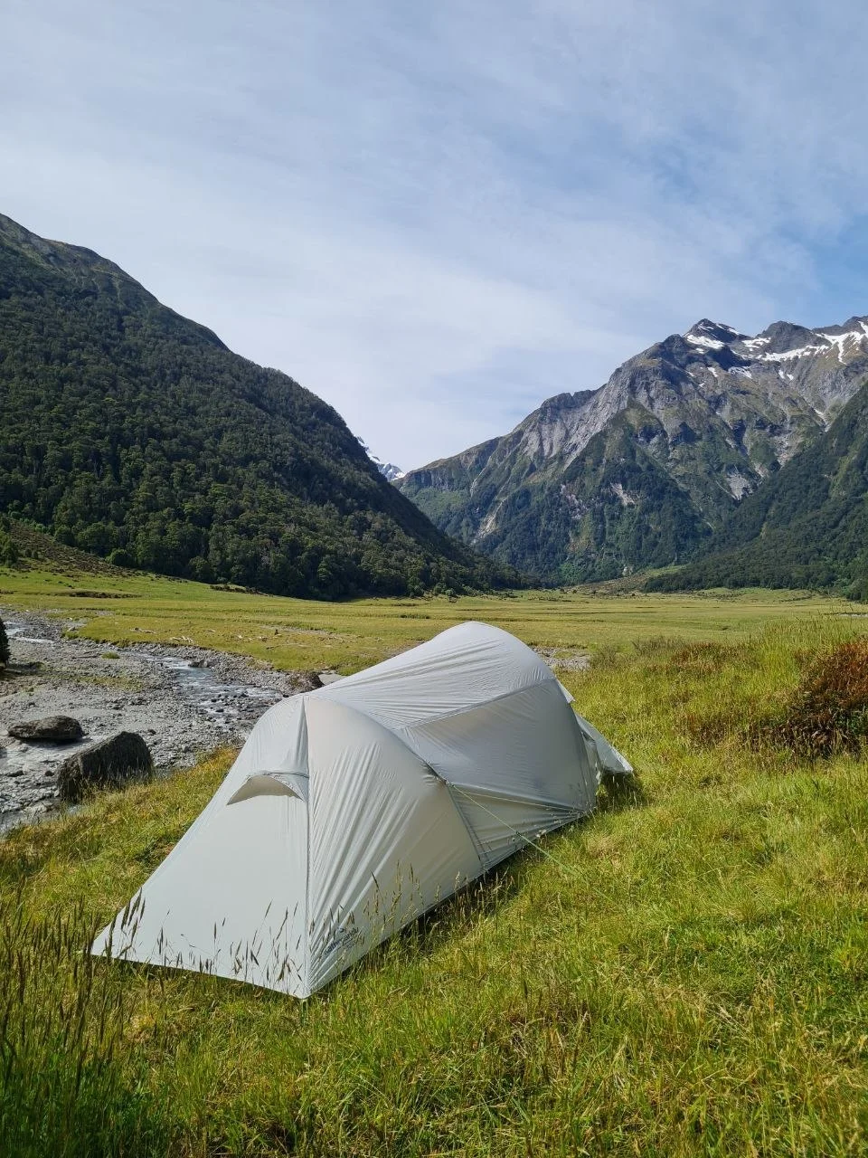 Tent pitched beside a clear stream in the upper valley, snow-patched mountains behind.
