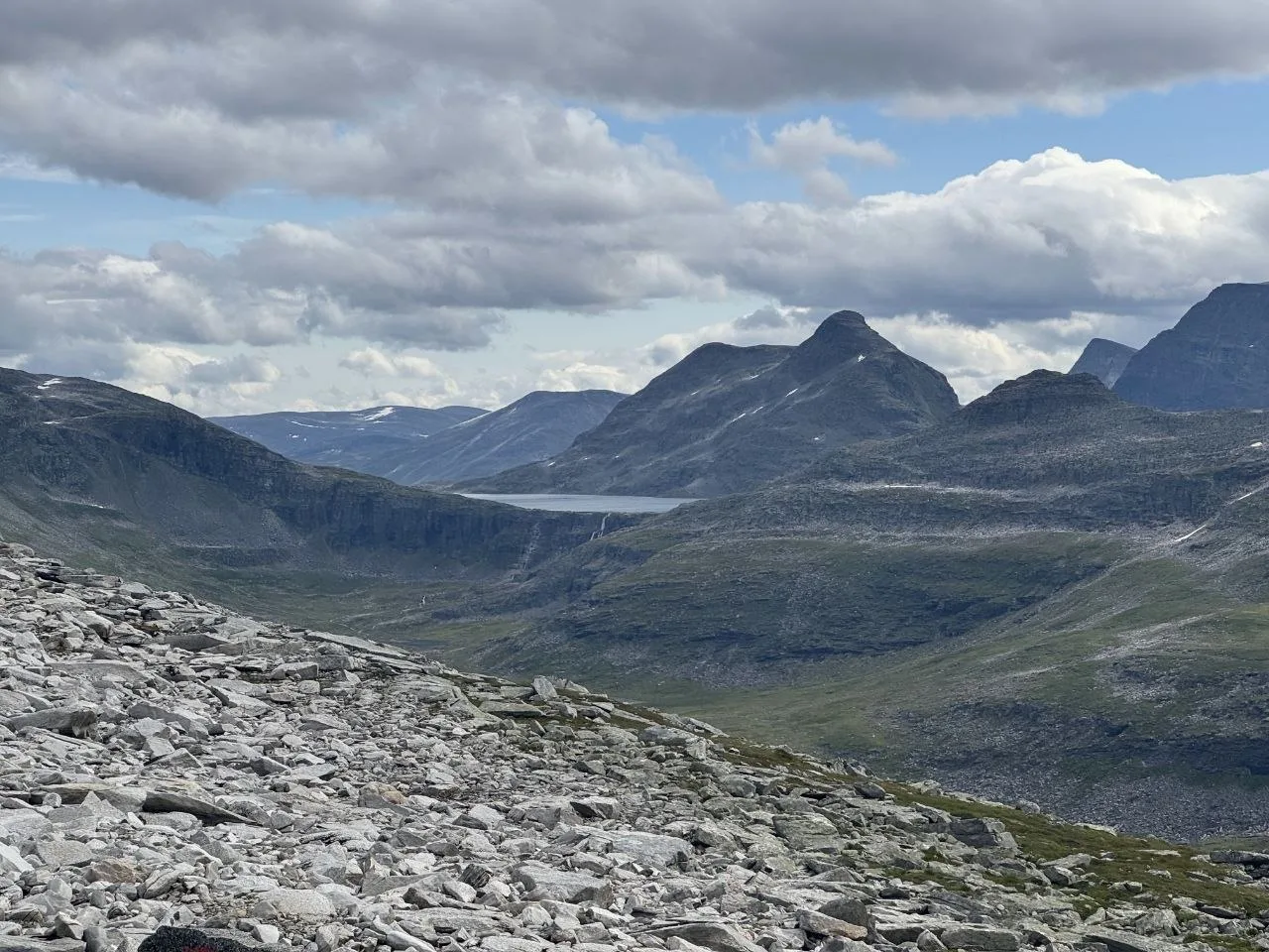 Rock-strewn mountain landscape with distant lake under cloudy sky